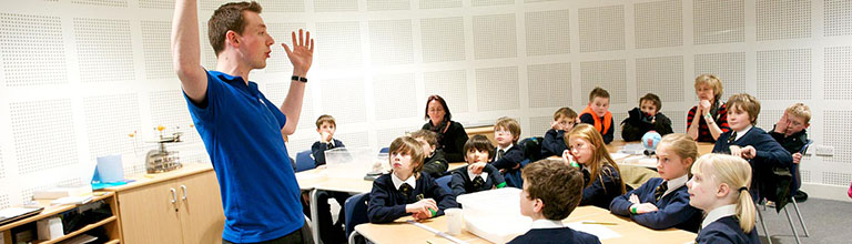 A teacher with arms raised in front of a class of young people