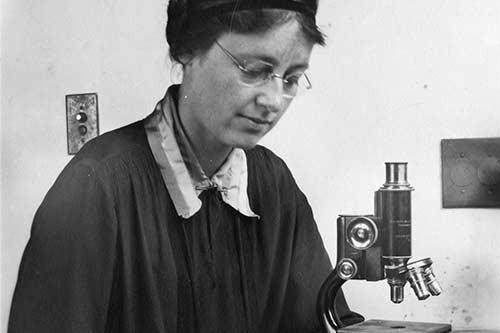 Old black and white profile of a lady sat next to a microscope. 