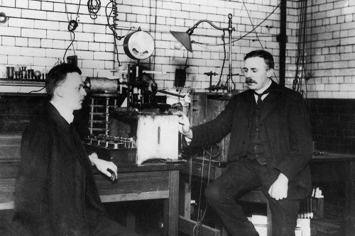 Old black and white photo of two men wearing suits and sitting  next to industrial machinery