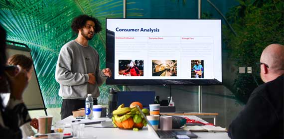 Man presenting to a team, stood next to a screen that has the words Consumer Analysis