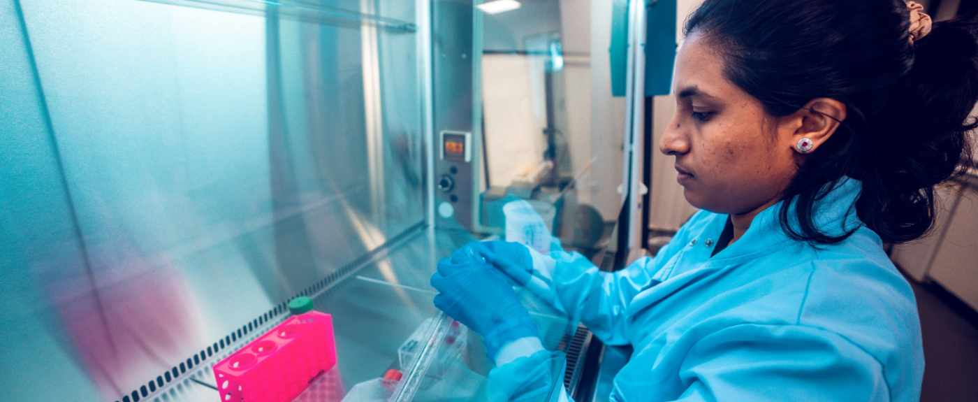 Female student in blue lab coat and blue gloves opens something behind a lab screen