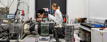 Two students in lab coats looking at electrical equipment