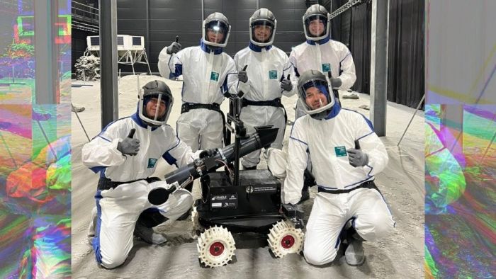 Group of people in protective suits posing with a rover vehicle in a simulated space environment.