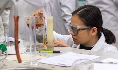 A student, wearing a white lab coat and safety googles, pours liquid from a beaker