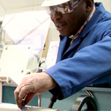 Male wearing safety goggles and blue lab coat reaching with hand