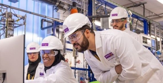 Researchers wearing lab coats, hard hats and safety goggles working together at a computer in a lab.
