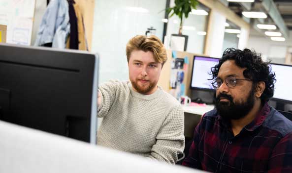 Two males sat looking at a computer screen while one of them is pointing to it