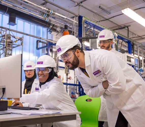 Researchers wearing lab coats, hard hats and safety goggles working together at a computer in a lab.