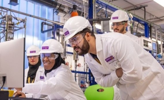 Researchers wearing lab coats, hard hats and safety goggles working together at a computer in a lab.