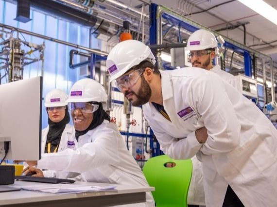 Researchers wearing lab coats, hard hats and safety goggles working together at a computer in a lab.