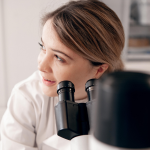 A CDT researcher in a white lab coat looking up from using a microscope. The microscope partially obscures their face.