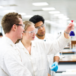 Three University of Manchester students in white lab coats and protective eyewear observe a beaker of blue solution in a Manchester laboratory.