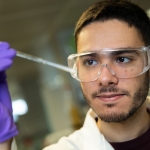 A PhD researcher at The University of Manchester in a white lab coat, protective glasses and purple gloves observing a pipet of a solution mid-experiment.