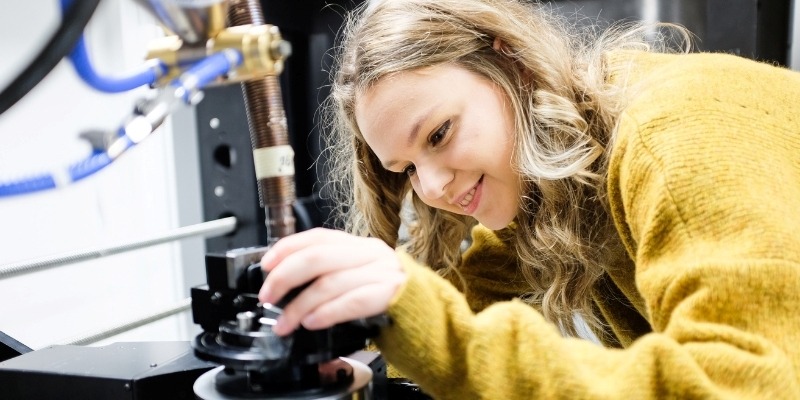 A maths student in a yellow jumper adjusts a component on a precise scientific instrument.