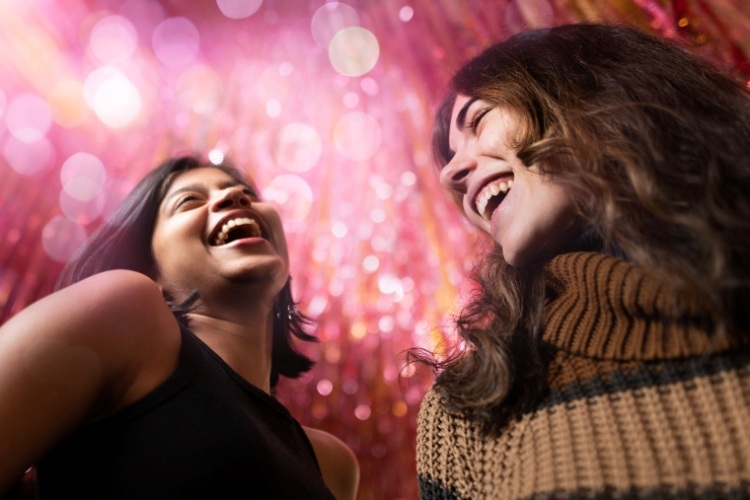 Two students laughing in front of a sparkling, blurred pink background.