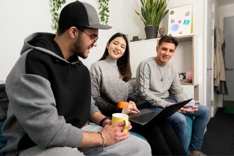 Students in uni accommodation talking around a laptop in a relaxed, upbeat setting.
