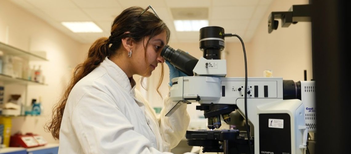 Deepanshi, wearing a white lab coat and blue gloves, looks through a microscope in a laboratory.