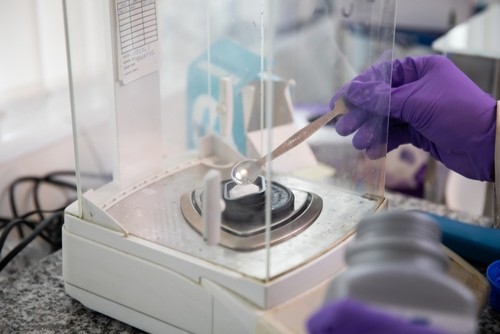 A person wearing purple nitrile gloves carefully placing a white powder onto a digital lab balance using a metal spatula.