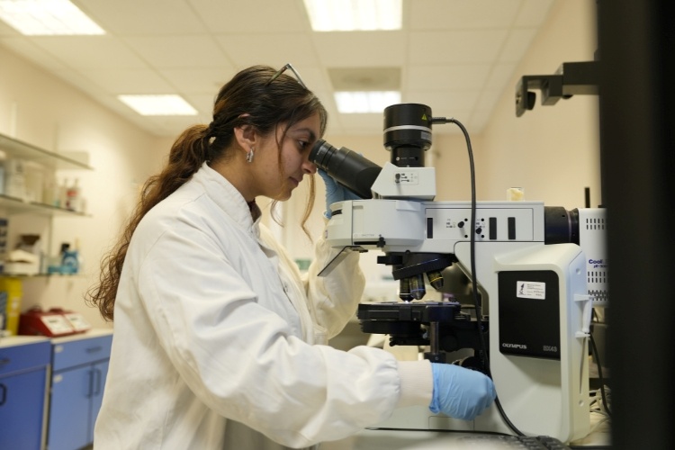 Deepanshi, wearing a white lab coat and blue gloves, looks through a microscope in a laboratory.