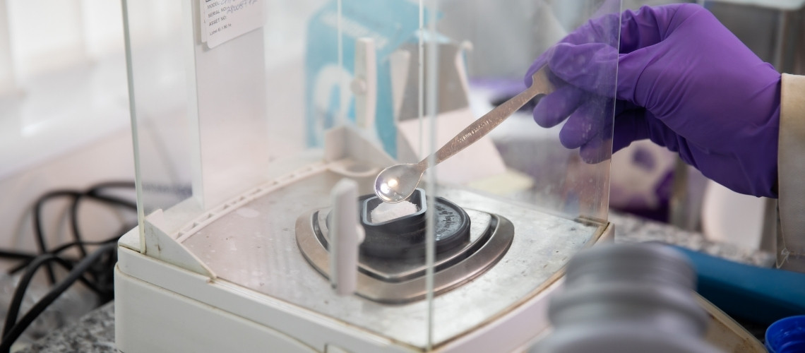 A person wearing purple nitrile gloves carefully placing a white powder onto a digital lab balance using a metal spatula.