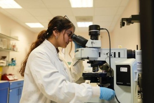 Deepanshi, wearing a white lab coat and blue gloves, looks through a microscope in a laboratory.