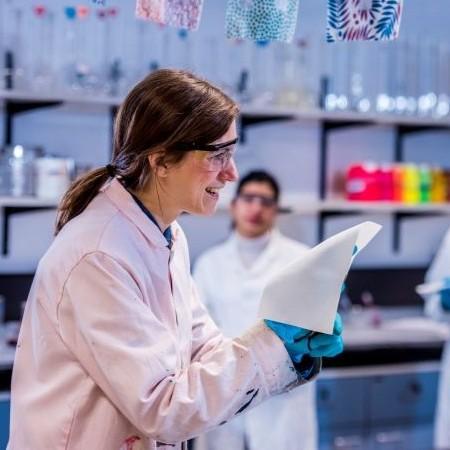 A smiling female scientist in a lab coat and safety goggles holds a piece of fabric while interacting with colleagues in a brightly lit laboratory.