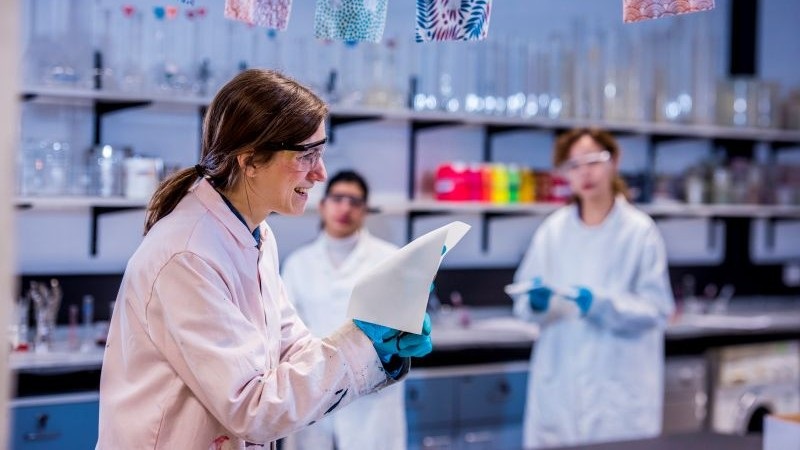 A smiling female scientist in a lab coat and safety goggles holds a piece of fabric while interacting with colleagues in a brightly lit laboratory.