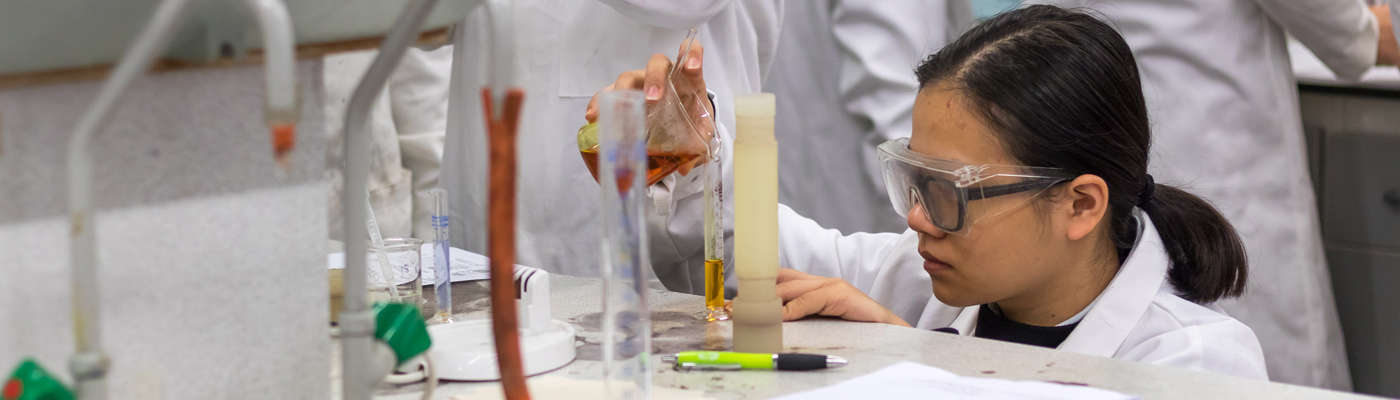 A female student, wearing a white lab coat and safety googles, pours liquid from a beaker